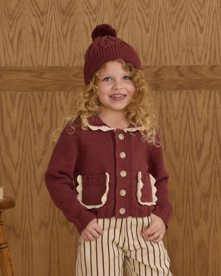 Child wearing a maroon knitted cardigan and hat against a wooden background